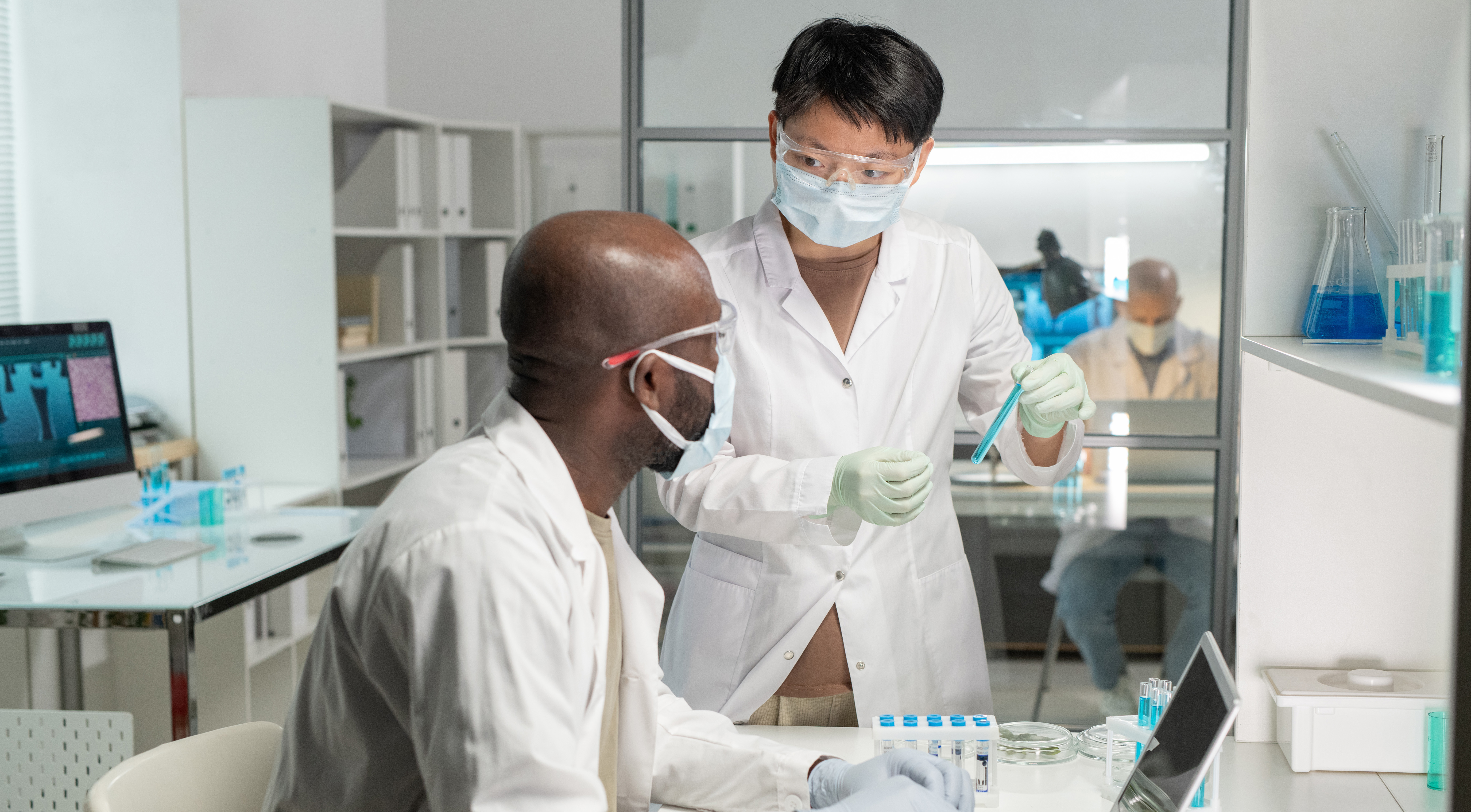 Researchers in lab coats, gloves, masks, and safety goggles review test samples and data beside a laptop in a laboratory.