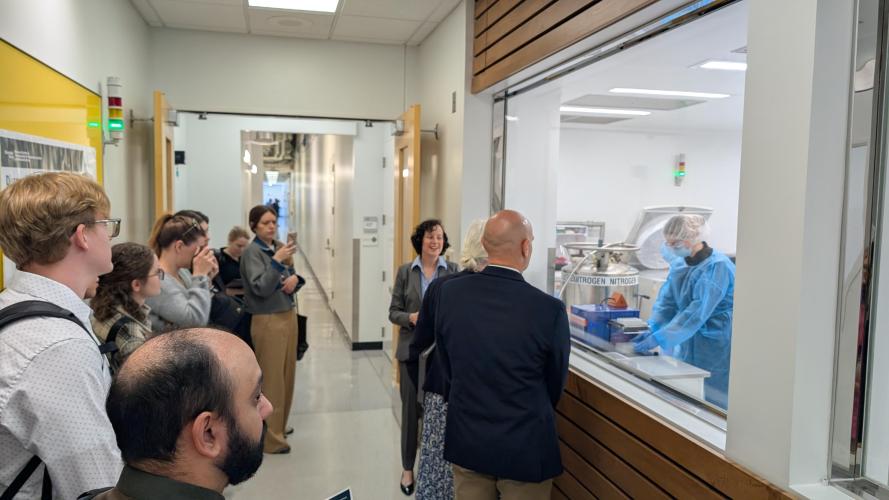 A group of people stand in a hallway in front of a large glass window overlooking researchers in a cleanroom. Two presenters speak to the group.