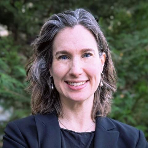 Headshot of Melissa Kemp, a white woman with brown and silver shoulder-length hair wearing a black blazer and blouse in front of green trees.